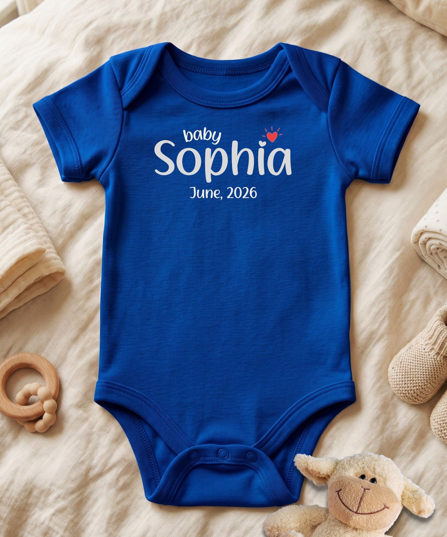 A royal blue short-sleeve baby bodysuit laid flat on a beige fabric surface, printed with the words 'baby Sophia June, 2026' in white, plus a small red heart and crown graphic. A plush toy, wooden ring, and folded textiles are arranged around it as props.