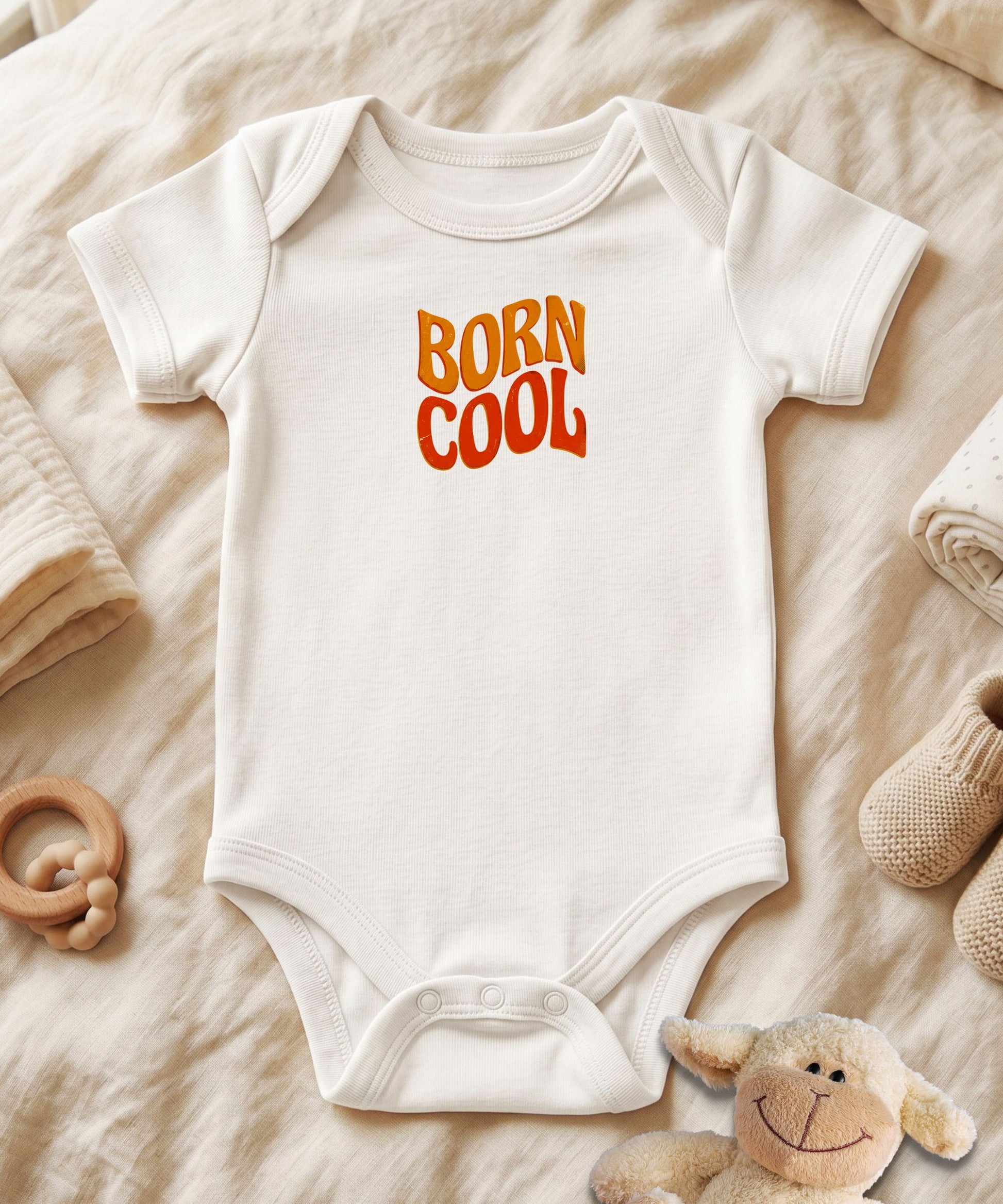 A white short-sleeve baby bodysuit laid flat on a beige fabric surface, featuring the words 'BORN COOL' in orange and yellow gradient lettering on the chest. Baby accessories, including a wooden ring rattle, a plush sheep toy, knit socks, and folded blankets, are arranged around it.